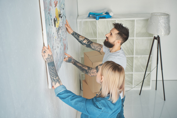 side view of young tattooed couple hanging painting on wall at new apartment - Photo, image