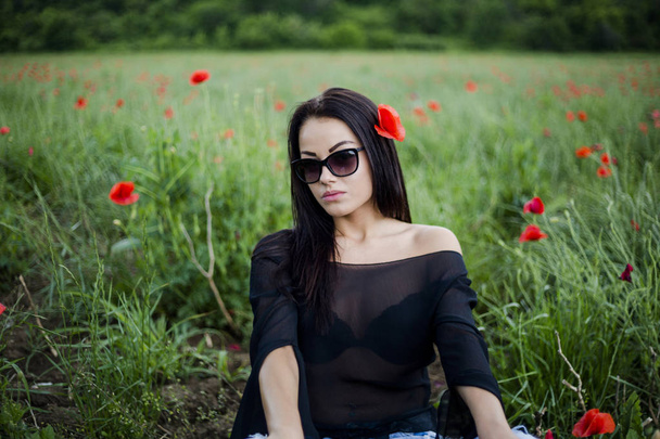 Krásná brunetka žena v sluneční brýle a stylové oblečení sedí mezi na červené vlčí máky na louce - Fotografie, Obrázek