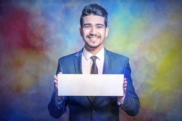 Young attractive man in a blue suit  holding a white empty sign on colorful abstract background - Photo, Image