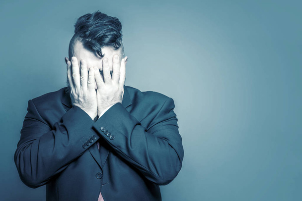 Adult man in a suit and a mohawk hairstyle covered his face on a blue background. Toned - Fotoğraf, Görsel