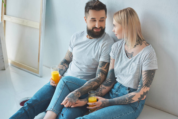 tattooed couple with glasses of juice holding hands while sitting on floor at new home - 写真・画像