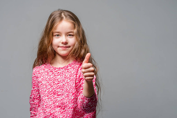 Cute little girl in a red dress shows thumb up on a gray background - Photo, image