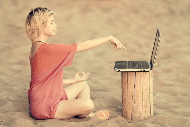 Girl blonde teenager in red blouse sitting on sand in lotus position and shows his hand on a laptop - Zdjęcie, obraz