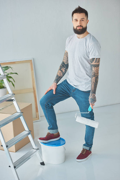 tattooed repairman with roll paint brush standing in new apartment with ladder and plastic bucket of paint - Φωτογραφία, εικόνα