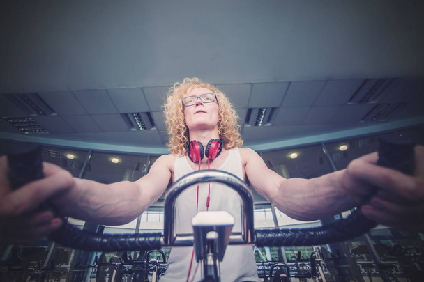 Homme blond bouclé avec des lunettes et des écouteurs rouges s'entraînant sur un vélo d'appartement dans la salle de gym
 - Photo, image