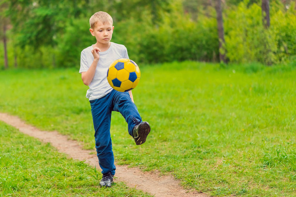 Blond boy in a white T-shirt walking along the path and kicking a soccer ball in the park. Toned - Foto, Imagem
