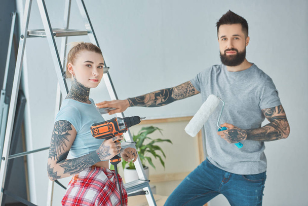 tattooed couple with screw gun and roll paint brush standing at ladder at new home - Photo, Image