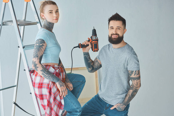tattooed couple with screw gun standing at ladder at new home - Photo, Image