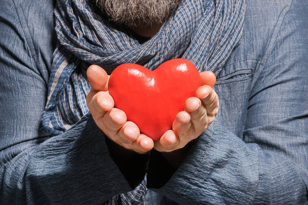 Adult bearded man in a blue jacket and scarf holding red heart - Foto, afbeelding