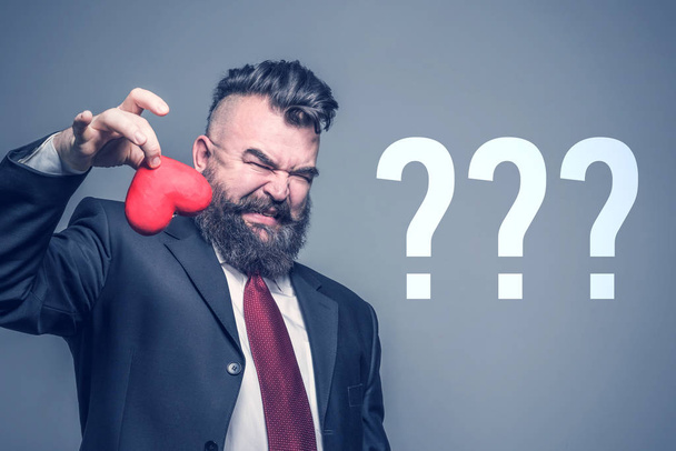 Adult bearded man in a black suit with disgust holding red heart on a gray background with the question marks - Foto, Bild
