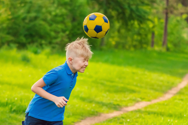 Blond boy in a white T-shirt boy beats his head a soccer ball in the park - Фото, изображение