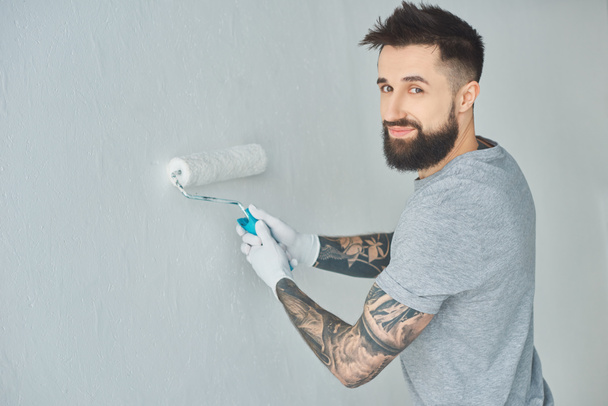 sonriente reparador tatuado con pincel de pintura en rollo mirando a la cámara mientras pinta la pared en un apartamento nuevo
 - Foto, Imagen