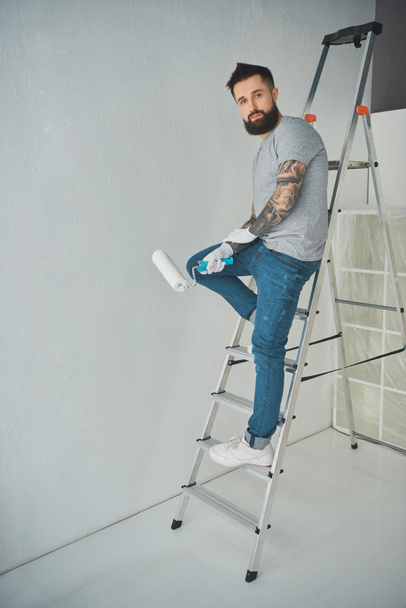 молодой татуированный ремонтник с кистью, стоящей на лестнице в новой квартире
 - Фото, изображение
