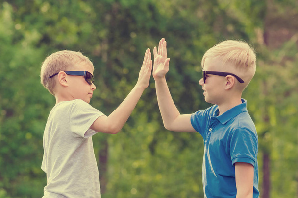 Two blond boy in blue and white T-shirts and sunglasses are giving high five in the park - Фото, зображення