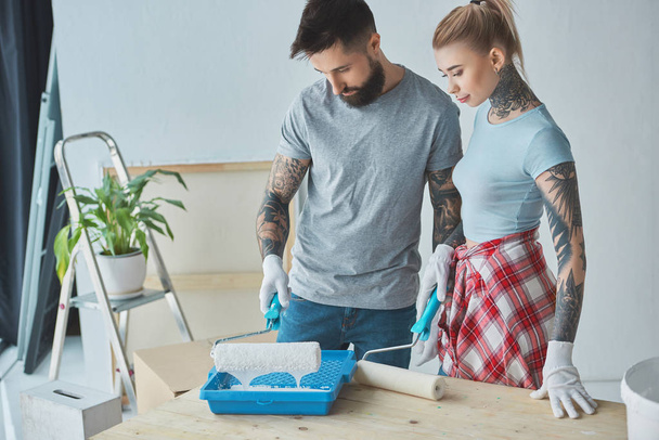 tattooed couple with roll paint brushes doing home repairment at new apartment - Foto, Bild