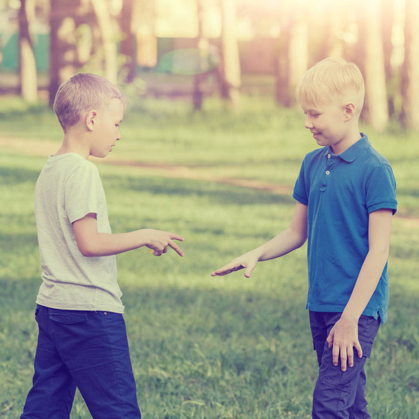 Two blond boys in white and blue T-shirts playing rock-paper-scissors in the summer park. Toned - Фото, изображение