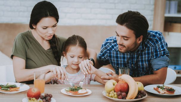 Teach Little Girl. Family Breakfast. Family at Table. Fruit on Plate. Sandwich on Plate. White Plate. Juice in Glass. Happy Family. Smiling People. Father and Mother Teach. White Interior. - Foto, immagini