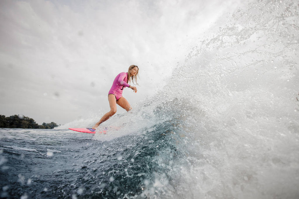 Jovem loira ativa wakesurfer vestido com maiô rosa descendo a onda azul salpicando em um dia nublado
 - Foto, Imagem