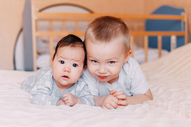 Söpö pieni neljän kuukauden ikäinen tyttö ja hänen isoveljensä, jotka leikkivät peiton alla kotona sängyssä makuuhuoneessa hymyillen iloisesti
 - Valokuva, kuva