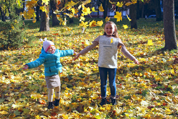 Young pretty sisters jump on yellow leaves in autumn park. Childs with autumn leaves. Little girls in autumn forest. Autumnal mood. Autumnal walk. Happy childhood. Good mood - Photo, Image