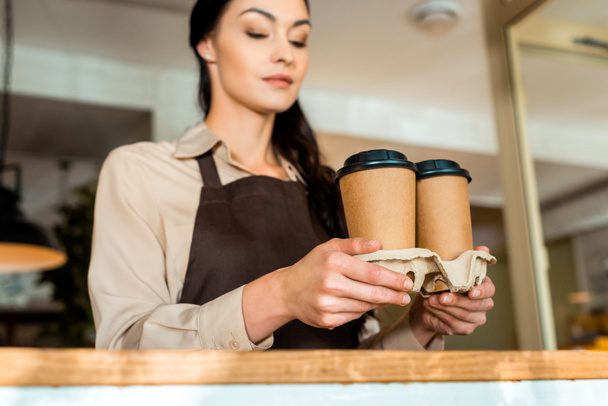 low angle view of attractive waitress in apron holding coffee in paper cups in cafe - Fotografie, Obrázek