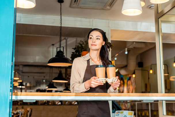 beautiful waitress in apron holding coffee in paper cups in cafe and looking away - 写真・画像