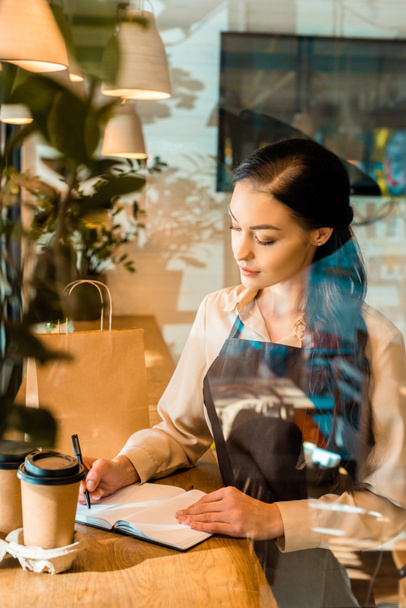 Blick durch das Glas einer attraktiven Kellnerin in Schürze, die im Café etwas zum Notizbuch schreibt - Foto, Bild