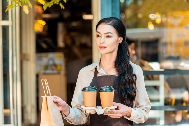 attractive waitress in apron holding two coffee to go and paper bag near cafe - Foto, imagen