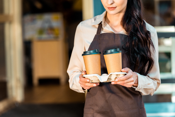 cropped image of waitress in apron holding two disposable coffee cups near cafe - Φωτογραφία, εικόνα