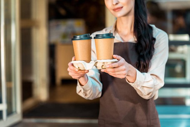 abgeschnittenes Bild einer Kellnerin in Schürze, die in der Nähe eines Cafés zwei Kaffees in Pappbechern hält - Foto, Bild