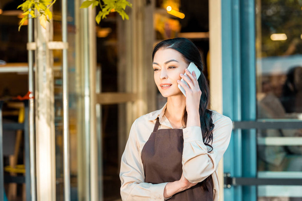 hermosa camarera en delantal hablando por teléfono inteligente y mirando lejos cerca de la cafetería
 - Foto, imagen