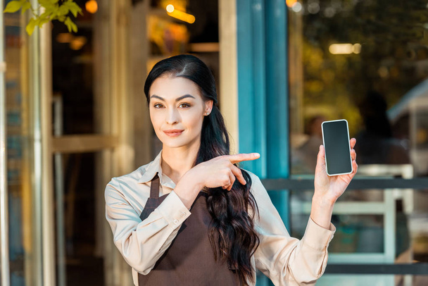 beautiful waitress in apron pointing on smartphone with blank screen near cafe - Zdjęcie, obraz