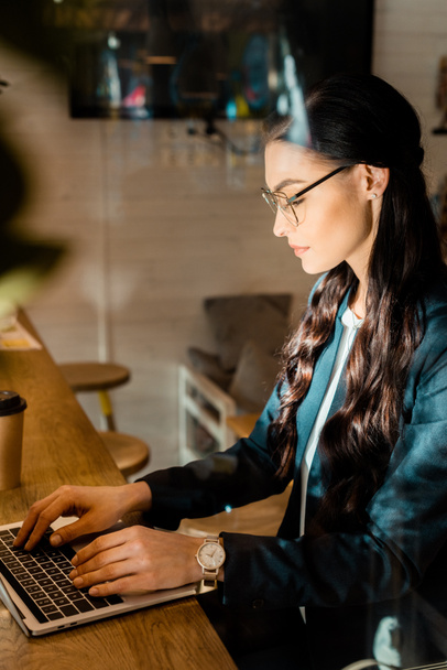 beautiful brunette teleworker in eyeglasses using laptop at restaurant - Photo, Image