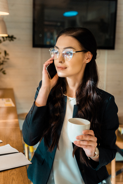 attractive brunette businesswoman in eyeglasses talking on smartphone and holding coffee cup in coffee shop - Фото, зображення