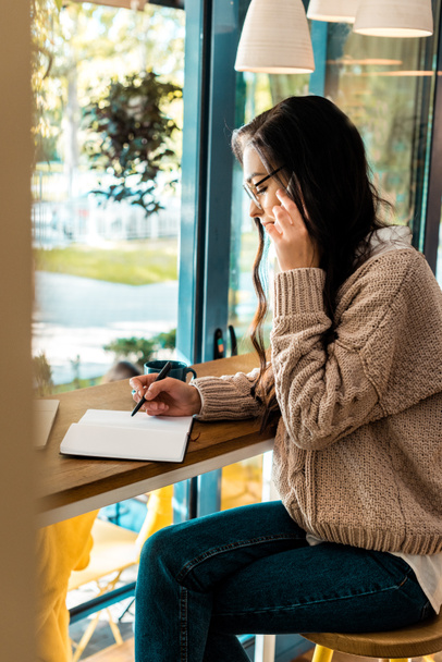 brunette femme pigiste parler sur smartphone et écrire dans un journal intime dans un café
  - Photo, image