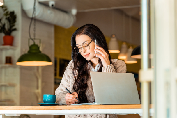 gyönyörű teleworker beszél a smartphone és írásban tervező kávézóban laptop - Fotó, kép