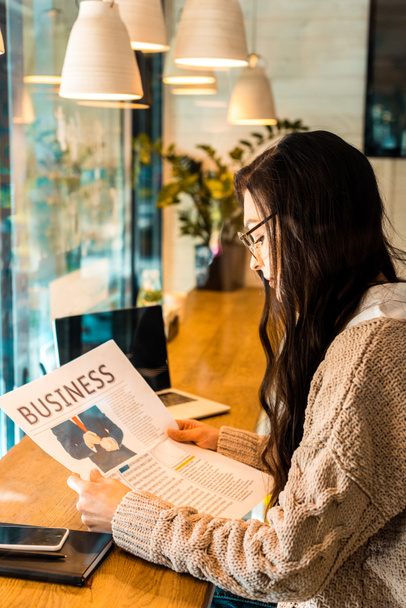 brunette woman reading business newspaper at cafe with laptop - Fotoğraf, Görsel