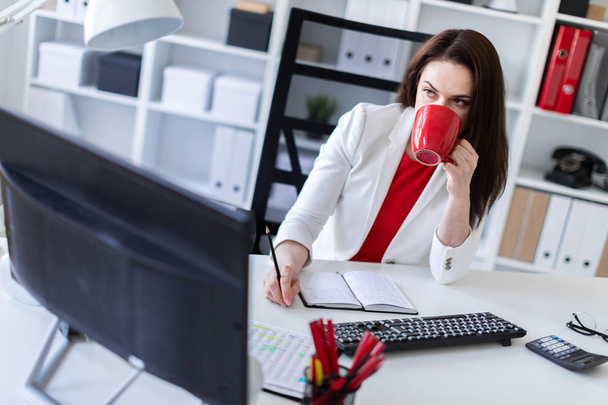joven mujer de negocios en la oficina cerca de la mesa, sosteniendo una taza roja
 - Foto, Imagen