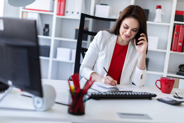Молода бізнес-леді сидить за столом біля комп'ютера в офісі, розмовляє по телефону
  - Фото, зображення