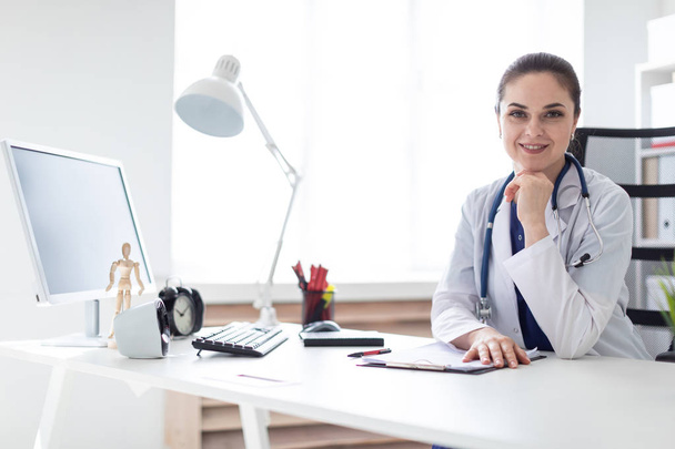Jeune médecin en blouse de laboratoire avec phonendoscope autour du cou assis à table au bureau
  - Photo, image