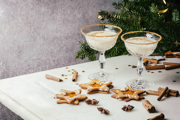 Cóctel de leche de Navidad de huevo, servido en dos vasos de cristal vintage con galletas de azúcar en forma de estrella de shortbread, palitos de canela, rama de abeto sobre mesa de mármol blanco
. - Foto, Imagen