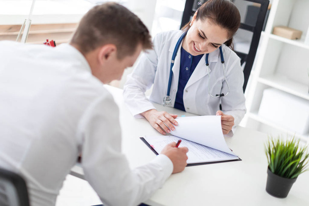 молодой пациент, сидящий с врачом за столом и подписывающий документы медицинского страхования в офисе
 - Фото, изображение