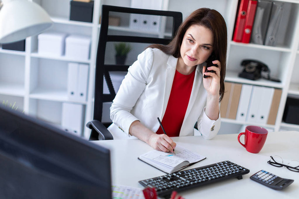 Молода бізнес-леді сидить за столом біля комп'ютера в офісі, розмовляє по телефону
  - Фото, зображення
