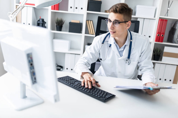 Молодой врач работает за компьютером, сидя с документами на рабочем месте в офисе
 - Фото, изображение