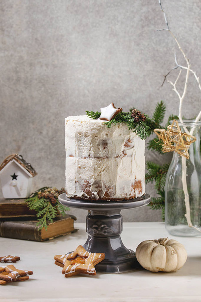 Autumn homemade white naked cake decorated by rated by star cookie and green thuja branches on cake stand fir tree, cookies, xmas decor above on white marble table. grey wall at background. - Fotó, kép