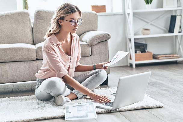 young woman in eyewear holding paper document while flooring at home with laptop on carpet  - Foto, imagen