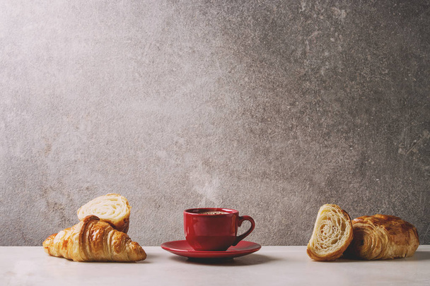 Fresh baked whole and sliced croissant with red cup of coffee espresso on white marble table with grey wall at background. Caffe breakfast - Valokuva, kuva