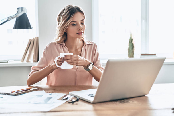 beautiful young woman in smart casual wear holding cup of coffee and looking at laptop while sitting at table with papers documents  - Photo, image