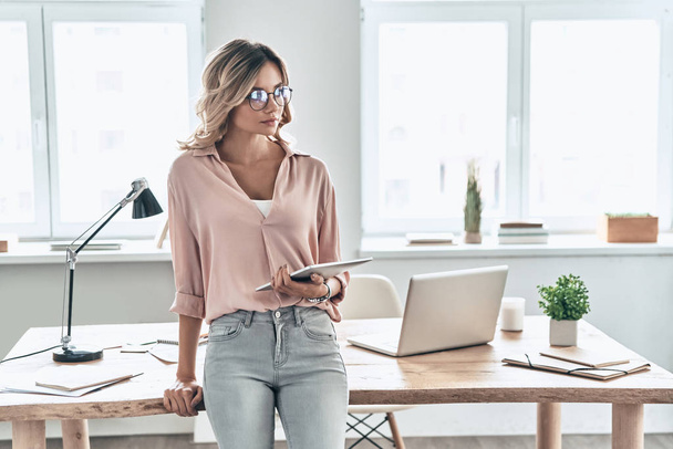 smart woman looking away while leaning on working desk in office, holding digital tablet  - Fotografie, Obrázek