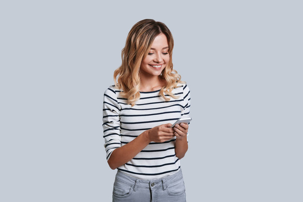 attractive young woman smiling and using smart phone while standing against grey background - Φωτογραφία, εικόνα